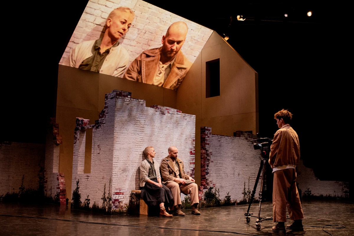 Actors on stage with a set of a house and a video projected above them