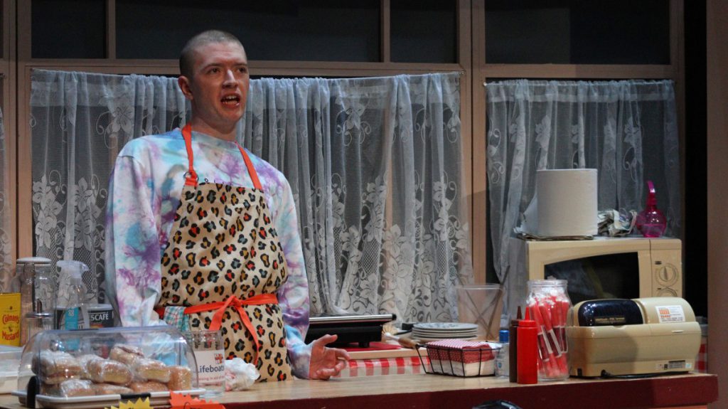 A young man stands behind a cafe counter on the set of the play Time and Tide